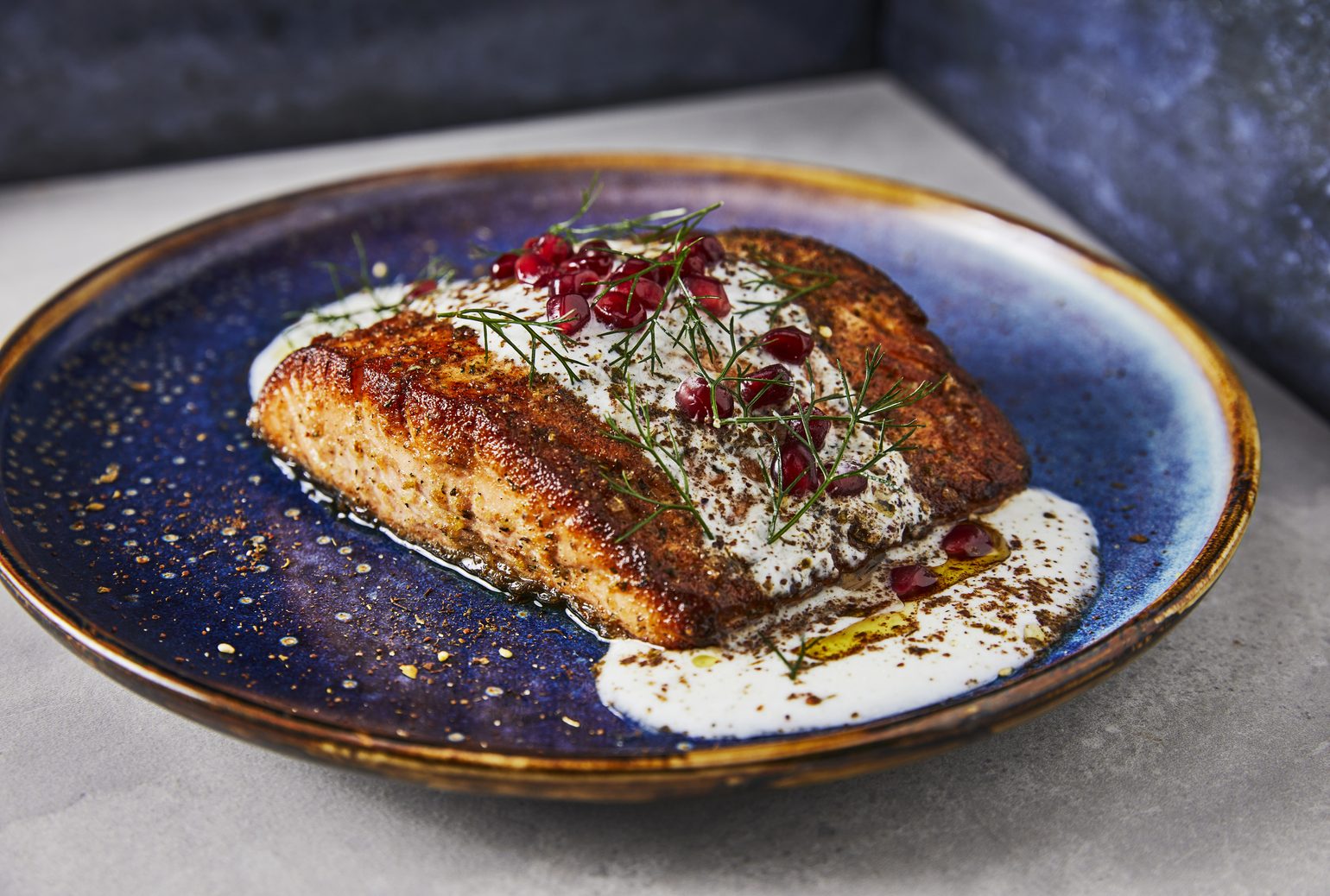 Pan-seared salmon with a pomegranate and dill garnish over a white sauce, on a speckled blue plate