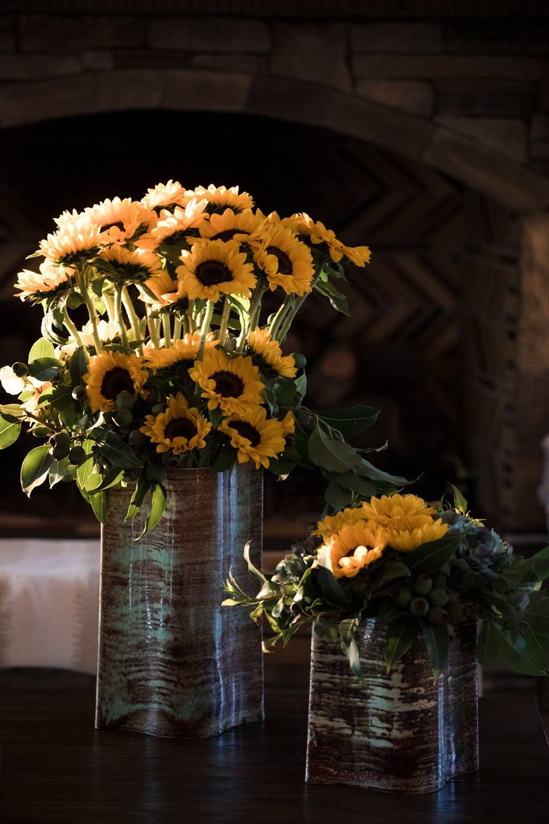 Sunflowers arranged in two bark-wrapped vessels on a pale cloth in front of a stone hearth