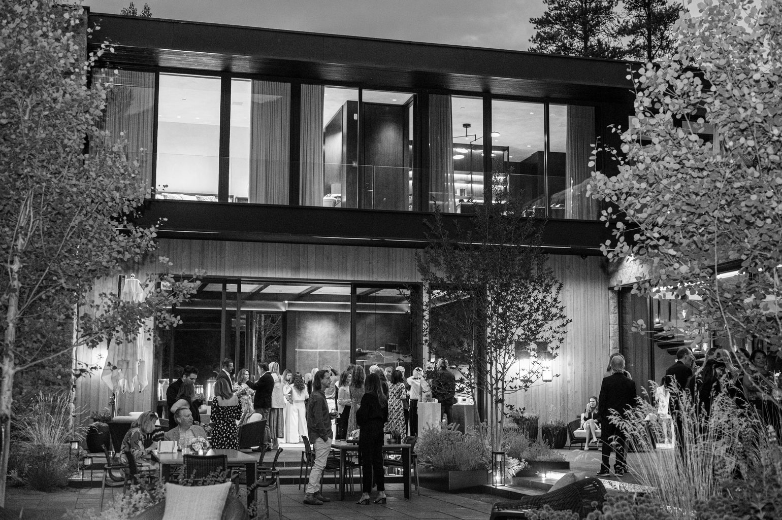 Guests at an evening party on the terrace of a glass-walled museum, in black and white.