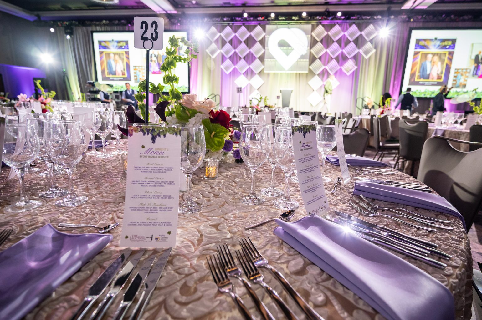 A gala table set with a printed menu by Chef Michelle Bernstein, lavender napkins and glassware, with a ballroom of round tables and a stage behind.