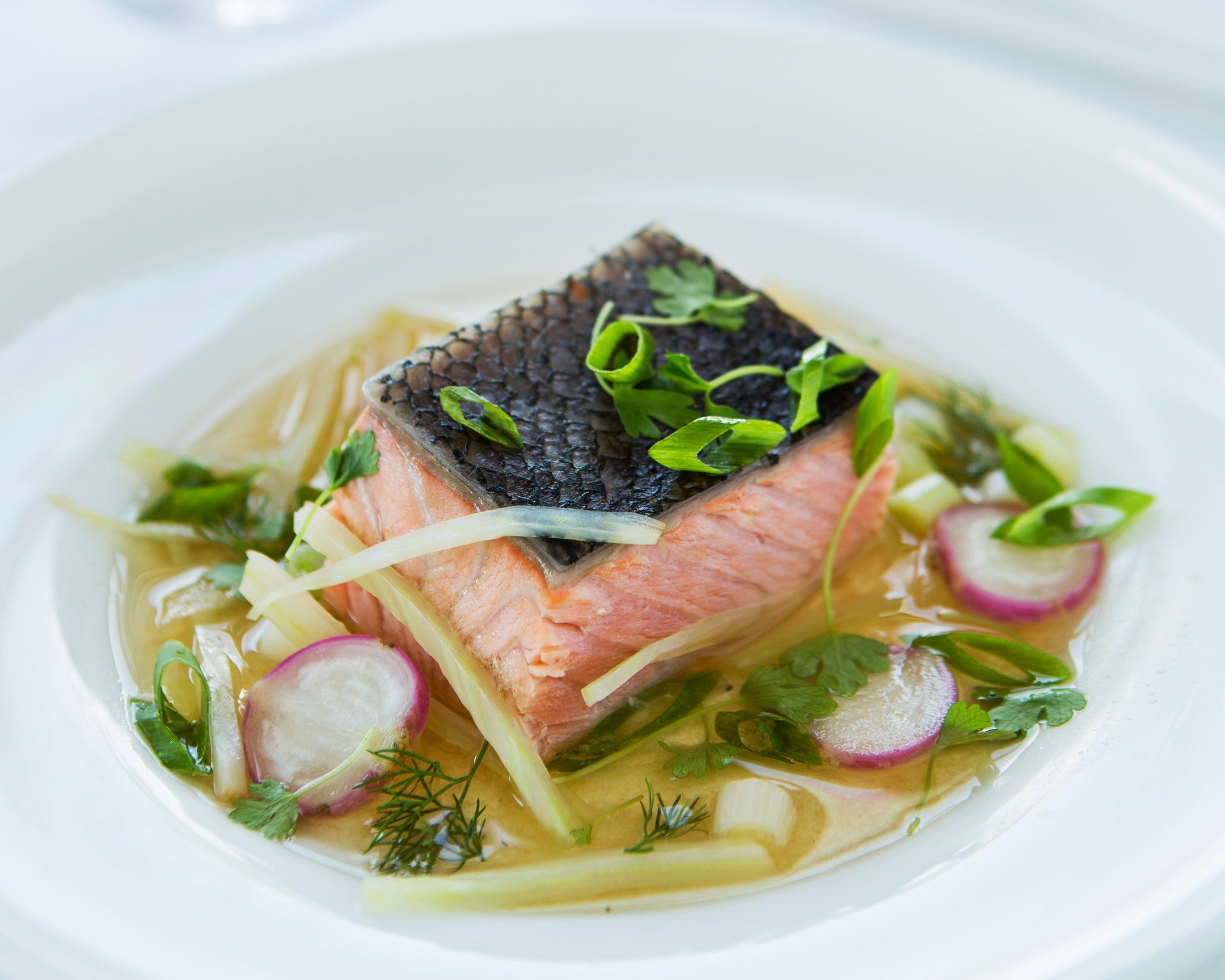 Poached salmon in a clear dashi broth with shaved radish and scallion, in a wide white bowl.