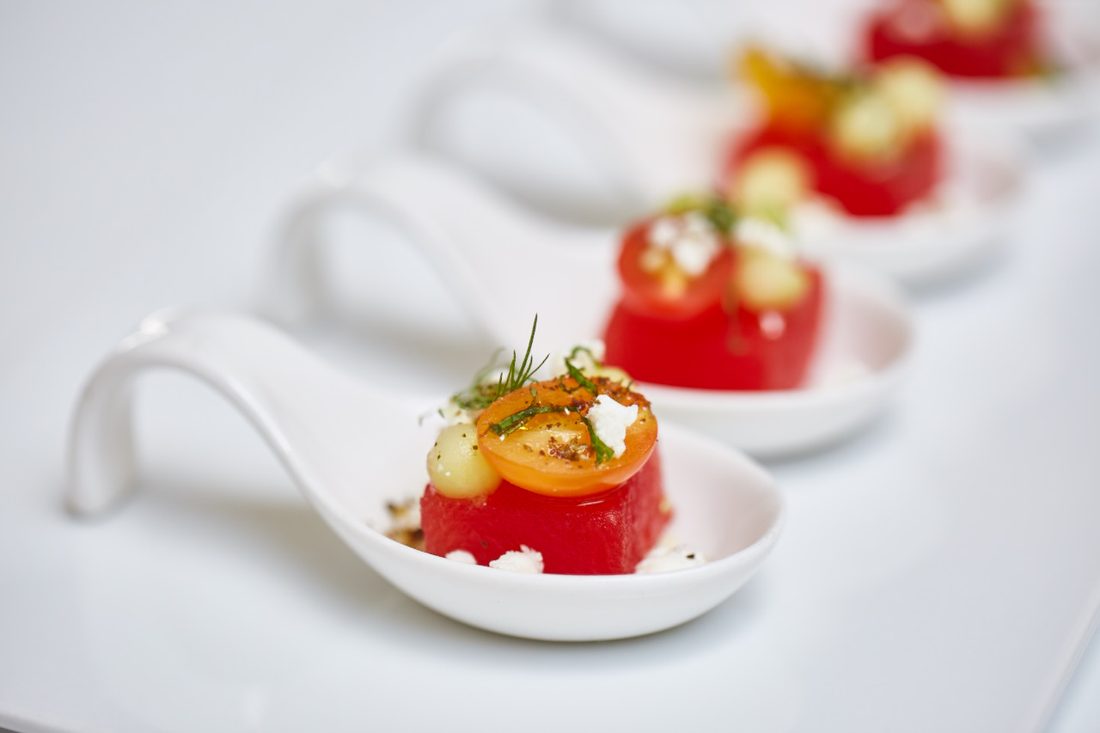 Watermelon canapés with feta and dill on white porcelain spoons, set out in a row.