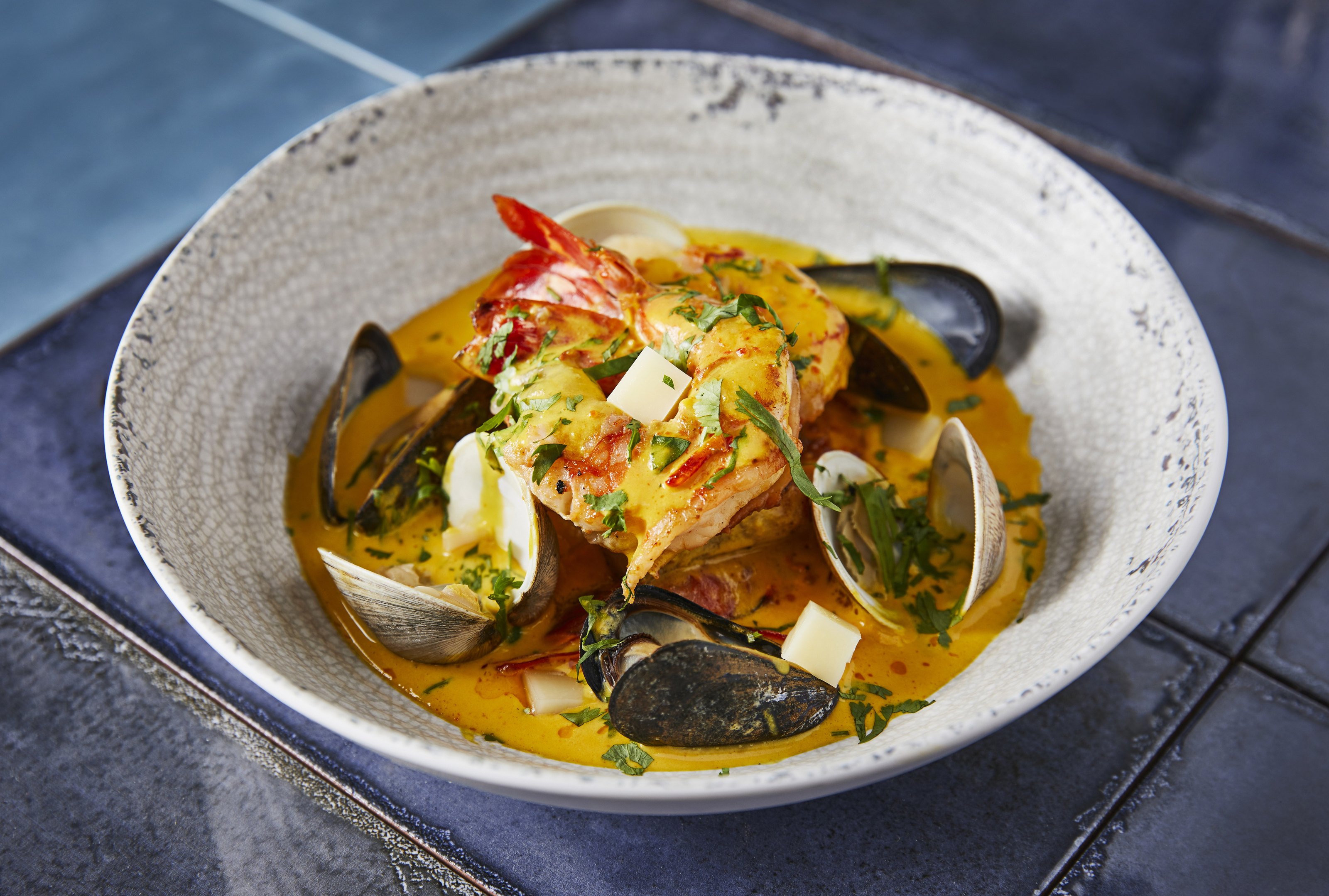 Mussels and lobster in a saffron broth, in a shallow bowl on blue tile.