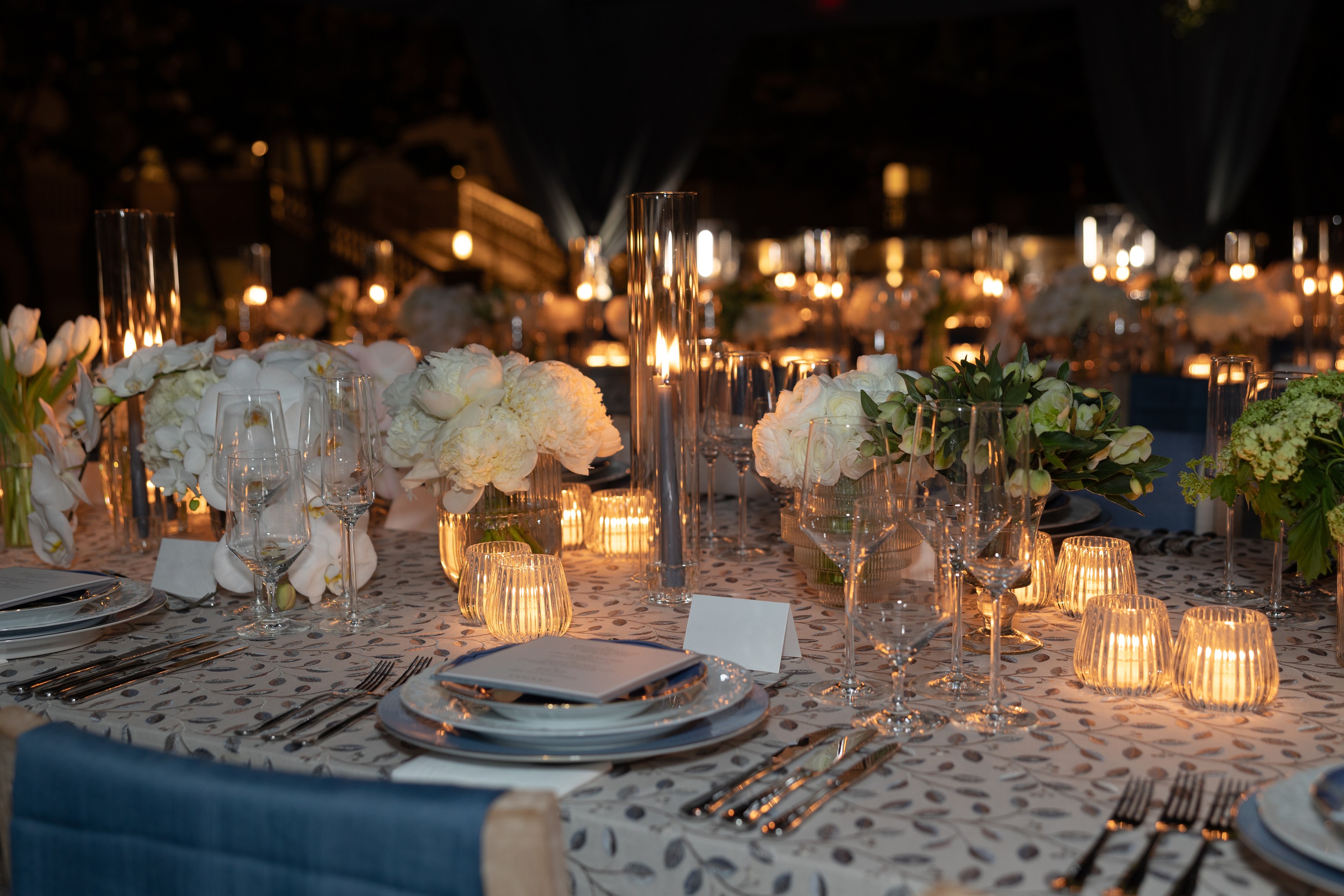 A candlelit round table set with white peonies and tulips, glass votives and printed menus on a leaf-patterned tablecloth