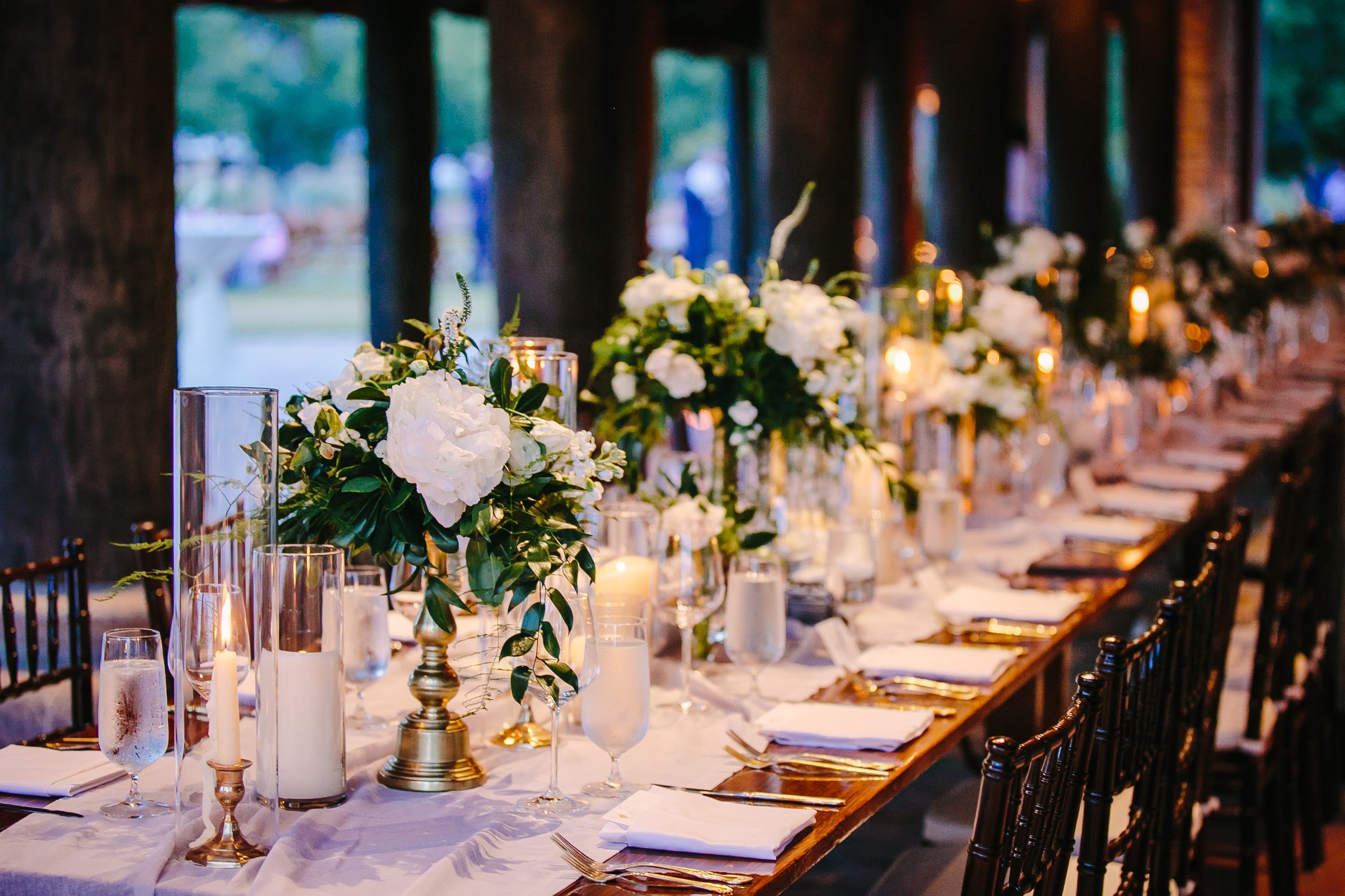 A long wedding table under candlelight, white peonies and roses in brass urns, gold flatware and napkins.